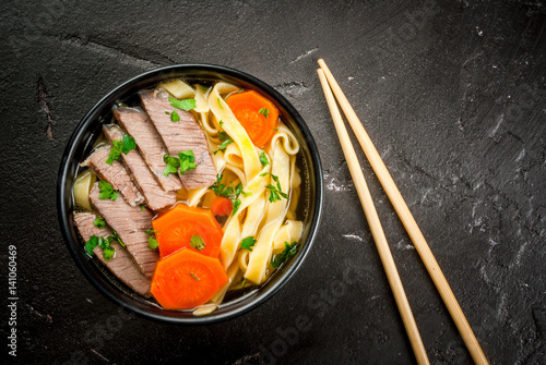 Beef soup with noodles and carrots in the Asian style. With chopsticks, on the black concrete table, top view, copy space