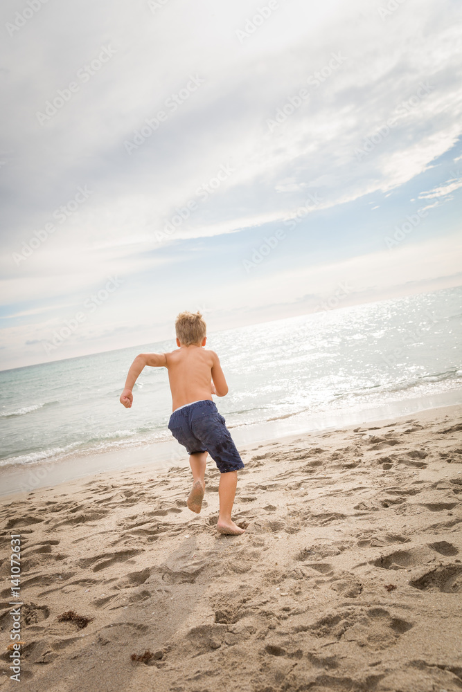 Cute kid boy running fast into the ocean on the sand beach. Clouds background and beautiful sea view. Active vacation by the sea. Sport.