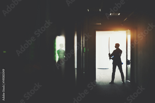 Silhouette of boss or businessman in formal suit standing in dark office corridor interior with reflections and green lights and holding katana sword like japanese samurai warrior ready to fight photo