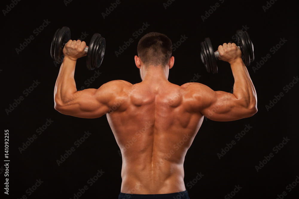 Muscular male model bodybuilder doing exercises with dumbbells, turned back. Isolated on black background with copyspace