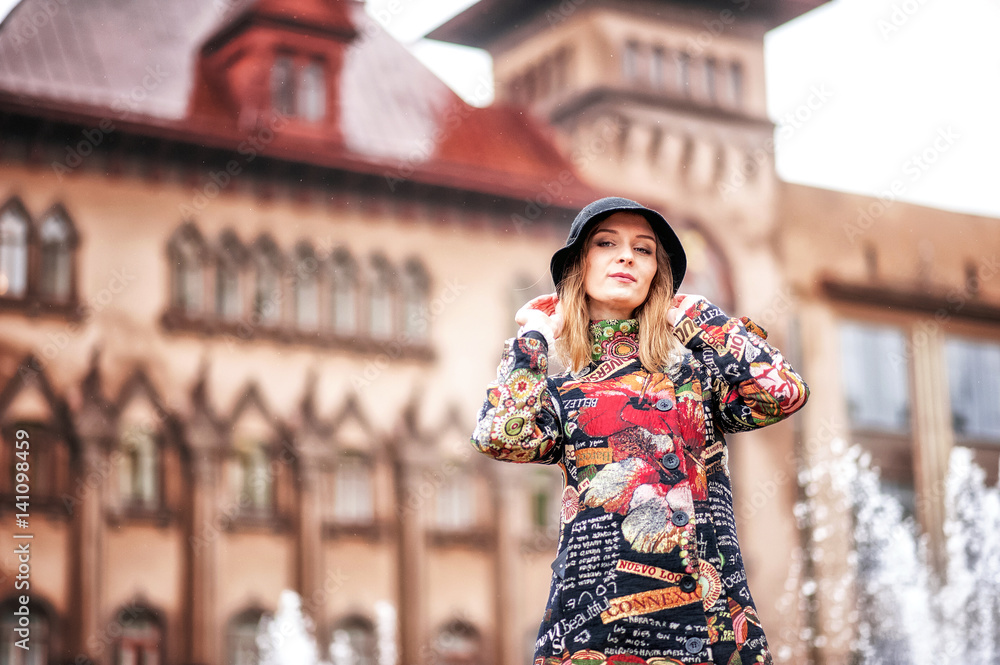 A beautiful girl in a bright multi-colored coat in rainy weather against a bright brick old castle or a house. Fountain in the background. Saratov, Russia.