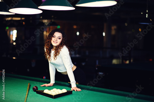 Young curly girl posed near billiard table. Sexy model at black mini mini skirt play russian snooker. Play game and fun concept.