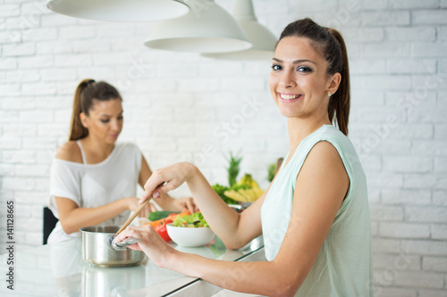 Young smiling woman cooking in kitchen. Her sister cutting vegetables in the background.