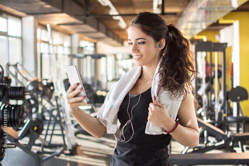 Fototapeta premium Attractive girl with earphones holding mobile phone to play favorite songs at gym.