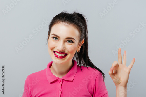 Beautiful young woman with curly long brown hair in pink mini dress posing with hand on hip and showing ok sign