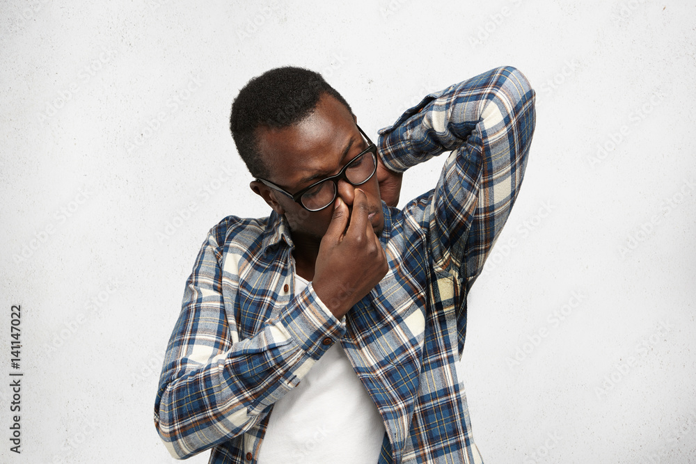Obraz premium Grumpy stinky young dark-skinned male in glasses and shirt raising elbow, sniffing sweaty armpit with disgusted expression, pinching nose, can't stand unpleasant smell of sweat, posing at white wall