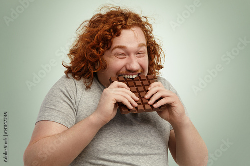 Fotografie Plump funny redhead young Caucasian male with curly hair biting large of chocola