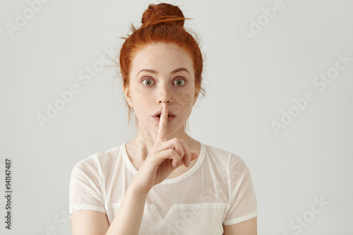 Portrait of beautiful young Caucasian woman with ginger hair bun holding index finger at lips, asking to keep silence or not tell anyone her secret, raising brows, saying 