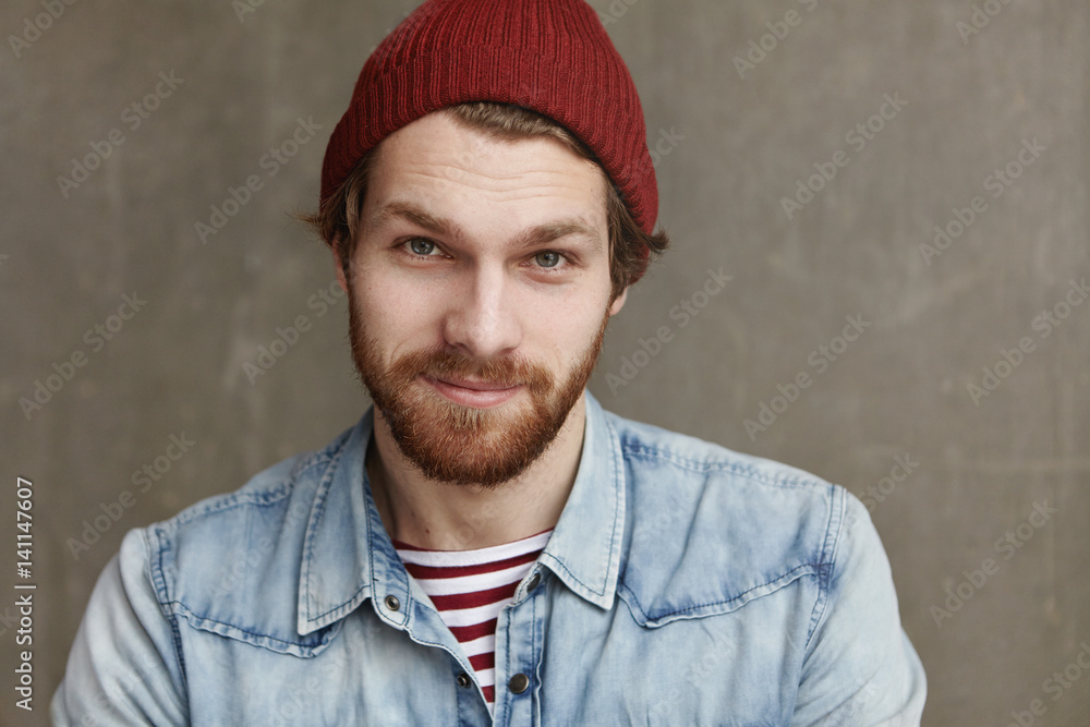 Close up shot of charismatic fashionable young European male with thick beard wearing trendy hat and denim jacket, staring at camera and smiling with happy cheerful look, enjoying free time at cafe Close up shot of charismatic fashionable young European male with thick beard wearing trendy hat and denim jacket, staring at camera and smiling with happy cheerful look, enjoying free time at cafe