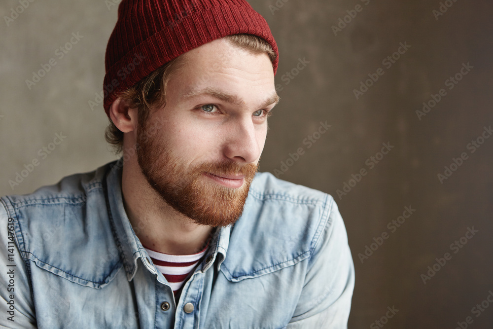 Indoor portrait of stylish good-looking young bearded hipster in trendy clothes with joyful mysterious smile, sitting at blank concrete copy space wall for your content, thinking about his girlfriend Indoor portrait of stylish good-looking young bearded hipster in trendy clothes with joyful mysterious smile, sitting at blank concrete copy space wall for your content, thinking about his girlfriend