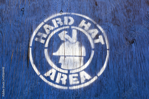 HARD HAT AREA construction site sign stenciled on blue wood panel.