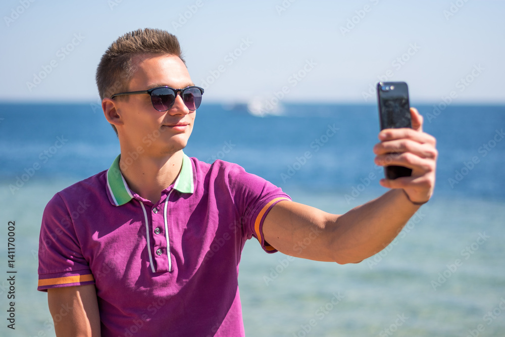 Man in sunglasses sits with a smartphone in the hands near the sea. Selfie.