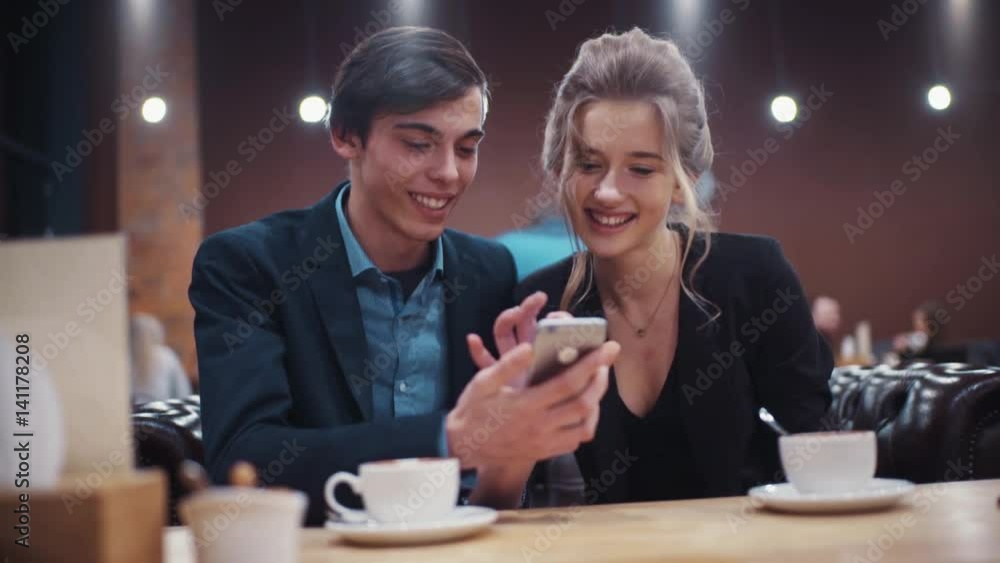 Young beautiful couple discussing cheerfully what they see on smartphone’s screen and drinking coffee on a date.