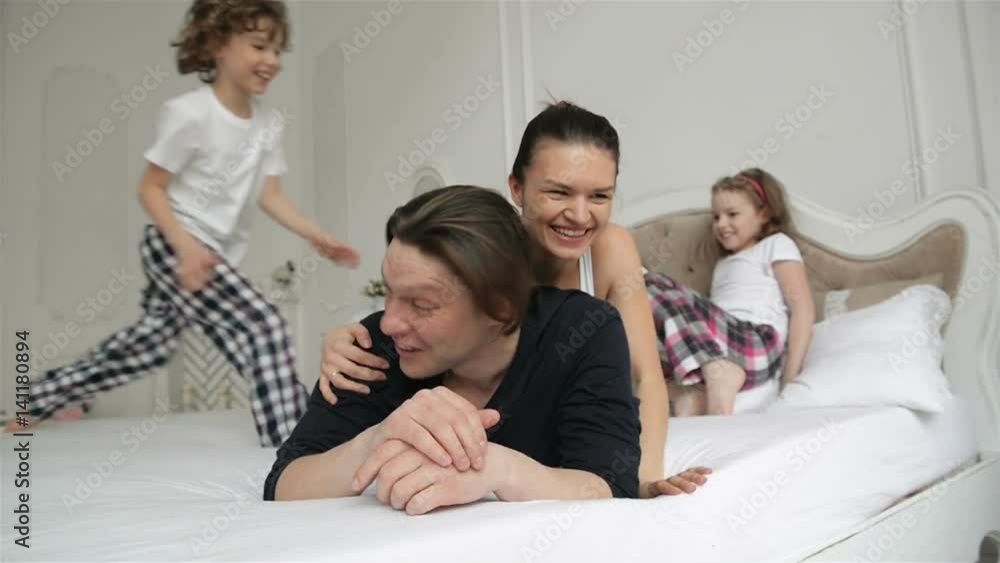Restless and Active Family Playing Together on a Bed at Home During Weeked. Young Parents with Charming Smiles are Spending Free Time with Their Son and Daughter in the Bedroom in the Morning. Restless and Active Family Playing Together on a Bed at Home During Weeked. Young Parents with Charming Smiles are Spending Free Time with Their Son and Daughter in the Bedroom in the Morning.