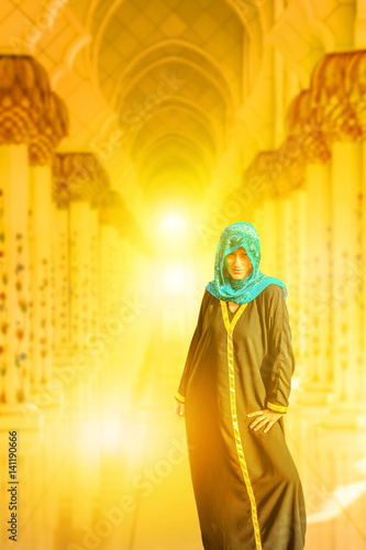 Woman with typical Arab clothes visiting the main attraction of Abu Dhabi and the largest mosque in UAE. Sheikh Zayed Grand Mosque at sunset. Travel Middle East concept. Defocused columns background.