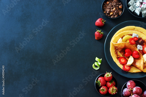 Healthy breakfast with homemade crepes with strawberries, grapes, dragon fruit, golden kiwi and honey over black background with a copy space. 