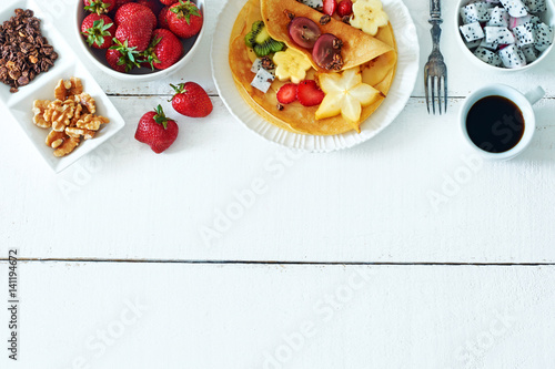 Top view of homemade crepes with exotic fruits, granola and black coffee over white background with a copy space. 