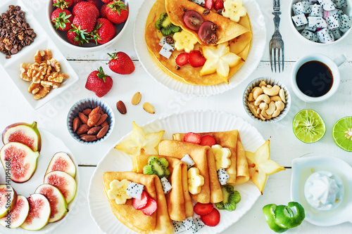 Healthy breakfast with thin pancakes, exotic fruits (starfruit, pitaya, banana, kiwi), strawberry, figs, granola, coffee and milk on a white table. 
