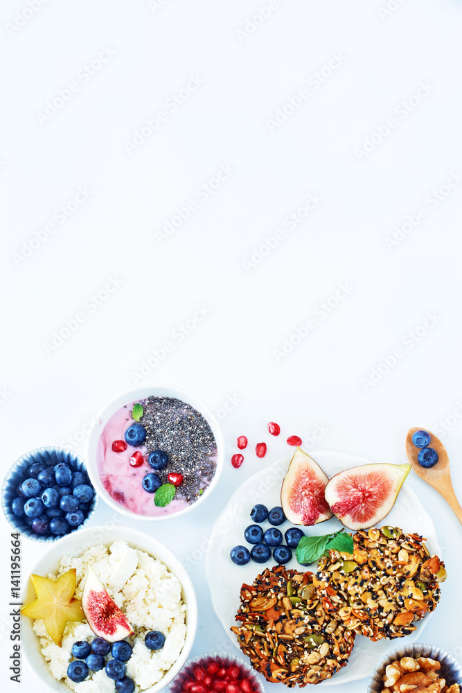 Vertical top view of healthy breakfast on a white table with a copy space. Multi-grain cookies, cottage cheese with starfruit, figs and blueberry, yoghurt with chia seeds, pomegranate and walnut. 