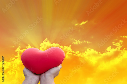 Hand gently hold red heart with love, careness and restpect on golden sky with clouds and sun ray background photo