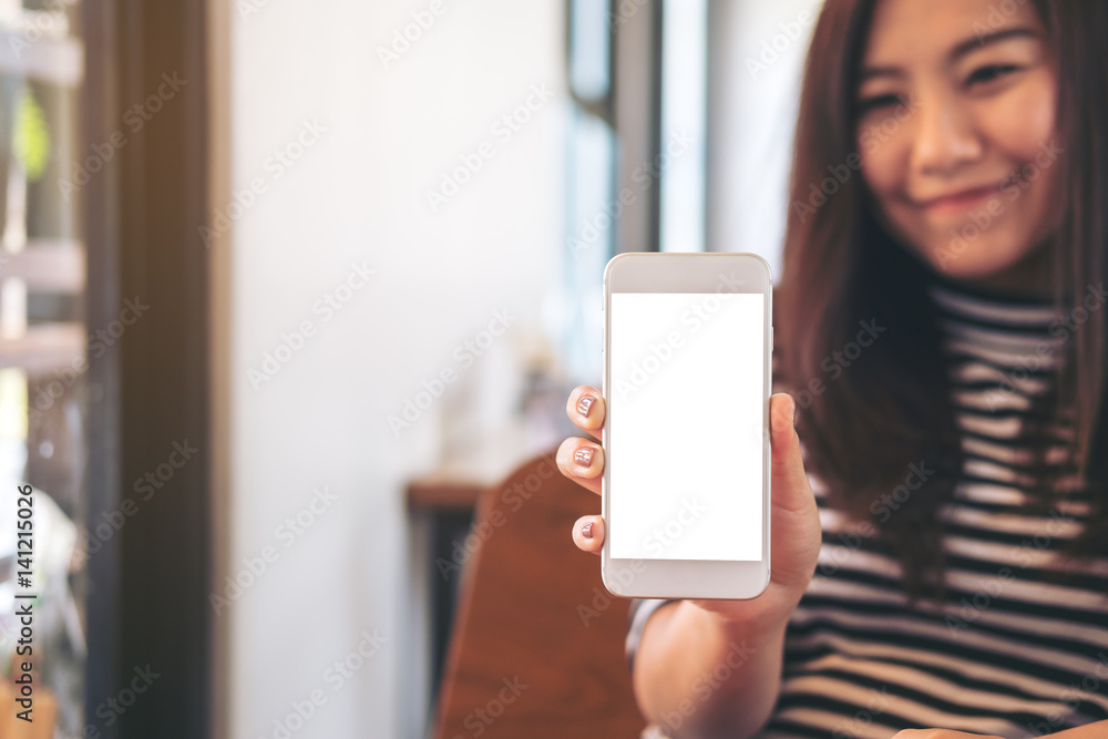 Mockup image of a beautiful woman holding and showing white mobile phone with blank white screen and smiley face in modern cafe