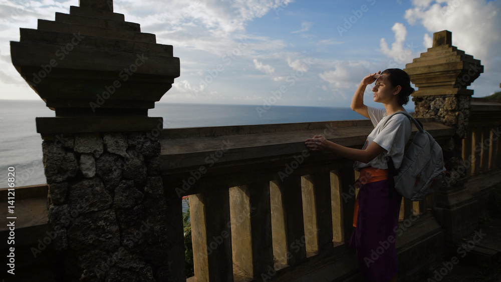 Girl with a backpack suited to a huge fence in front of a cliff and peering into the distance ocean shielding his eyes from the sun's light by hand.