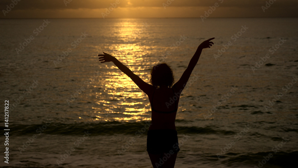 Slender girl slowly walks into the ocean at sunset to cool off. Silhouette of a young woman on a background of the sun reflections in the water, which raises hands up.