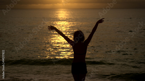 Slender girl slowly walks into the ocean at sunset to cool off. Silhouette of a young woman on a background of the sun reflections in the water  which raises hands up.