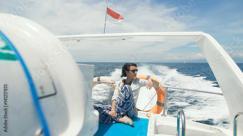 Cute girl sitting in the back of a motor boat enjoys walking on the ocean. Young woman in sunglasses look around and smiles. Tourist on a yacht while sailing.