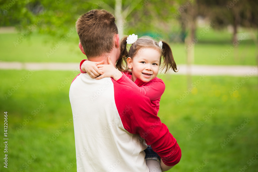 Fototapeta premium Young daddy holds her little daughter in hands while walking in city park. People enjoys warm weather on spring sunny day. Horizontal color photography.