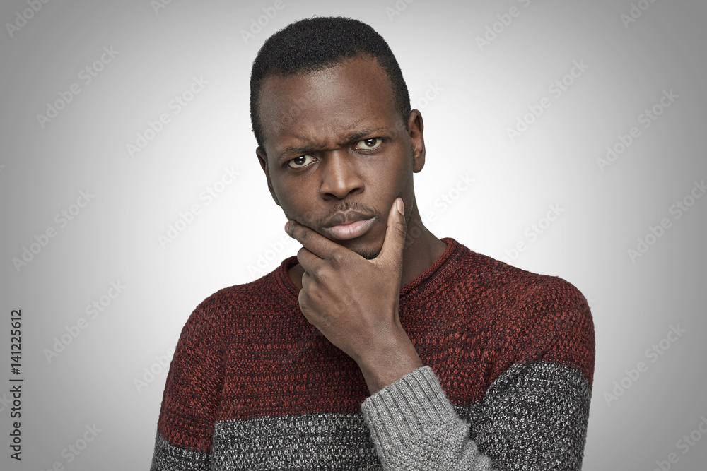 Close up portrait of serious puzzled African American male touching his chin, looking thoughtful and skeptical about something. Dark-skinned student deep in thoughts, hesitating to make decision Close up portrait of serious puzzled African American male touching his chin, looking thoughtful and skeptical about something. Dark-skinned student deep in thoughts, hesitating to make decision