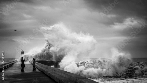 Crazy brave people on the pier near the lighthouse make photos during heavy storm against the backdrop of huge ocean waves. The Atlantic coast. Douro river mouth. Porto. Portugal. Europe.