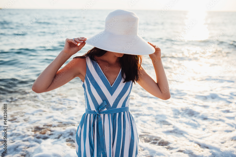 Attractive beautiful woman with white hat on head on sea sunset or sunrise background. Cover idea. Sexy female in dress, hide face with hat. Travel concept. Lifestyle fashion concept.