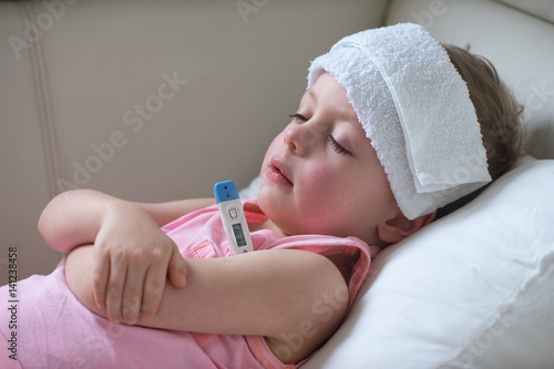  Sick child with high fever laying in bed and  holding thermometer.  Compress on forehead. photo
