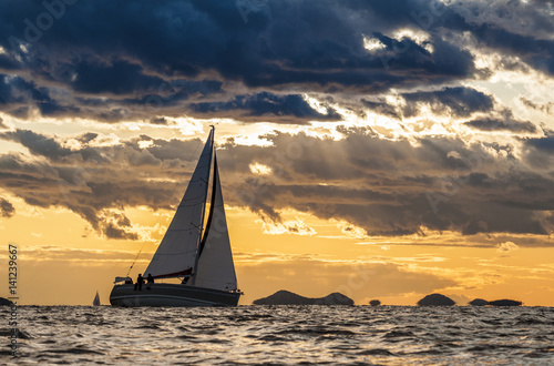 Sailing boat on open sea with dramatic clouds and small islands in background © Pavel