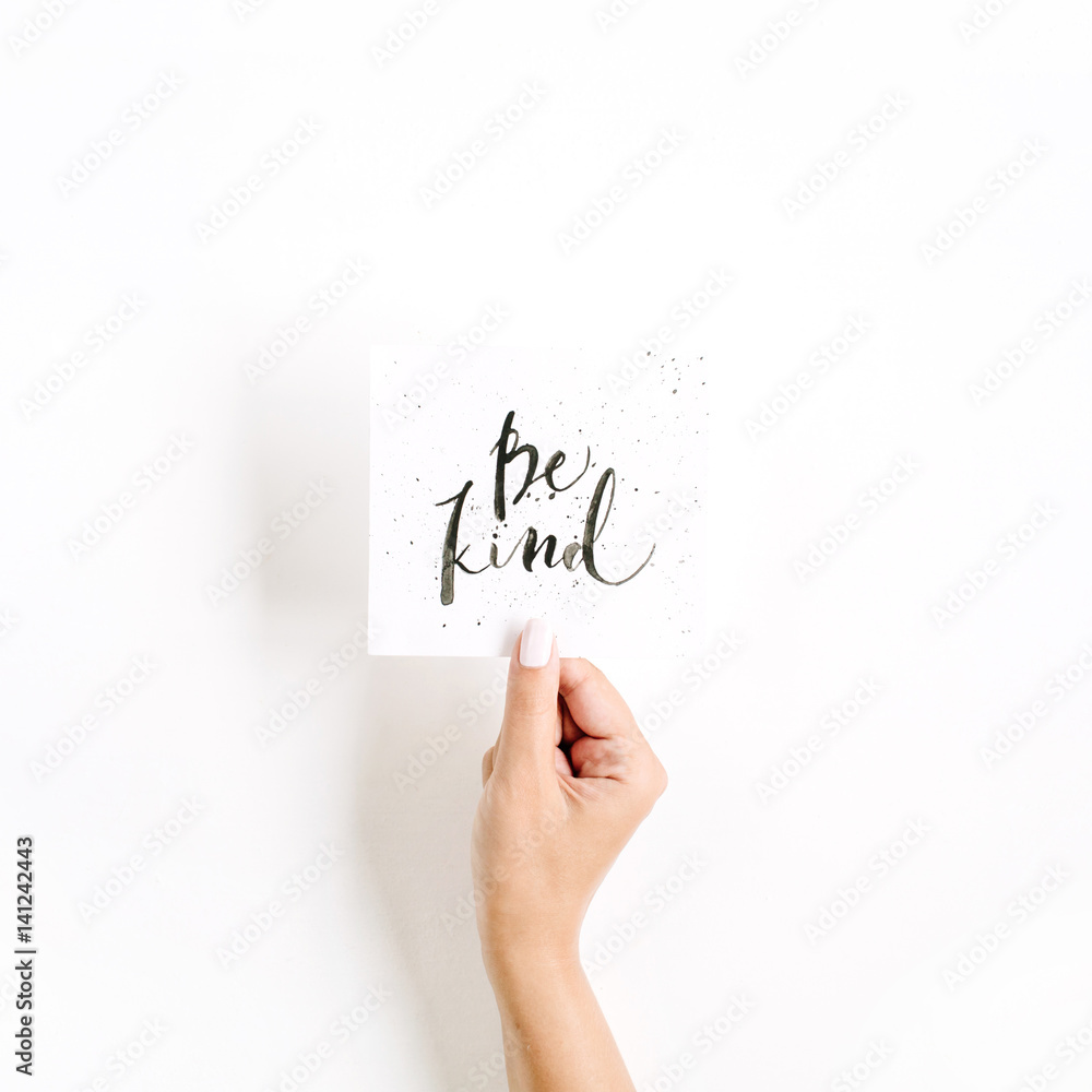 Minimal pale composition with girl's hand holding card with quote Be Kind written in calligraphic style on paper on white background. Flat lay, top view Minimal pale composition with girl's hand holding card with quote Be Kind written in calligraphic style on paper on white background. Flat lay, top view