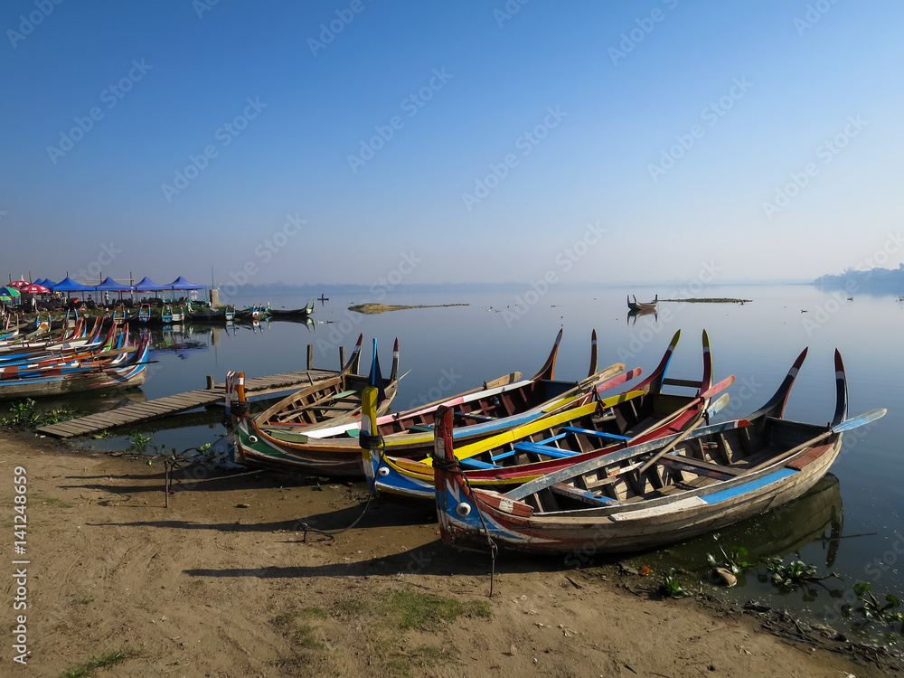 Colorful wooden boats on bank of peaceful still water lake with port, birds and clear blue sky background Colorful wooden boats on bank of peaceful still water lake with port, birds and clear blue sky background
