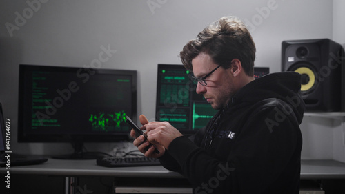 Adult serious man, looking like Hacker, with a small stubble on the face, points on the eyes, wearing a black hoodie with hood sitting in a dark room and produces count data with the help smartphone