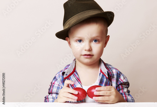 4 year old little and cute boy in a hat and shirt holding hands red Easter eggs, looks at viewer