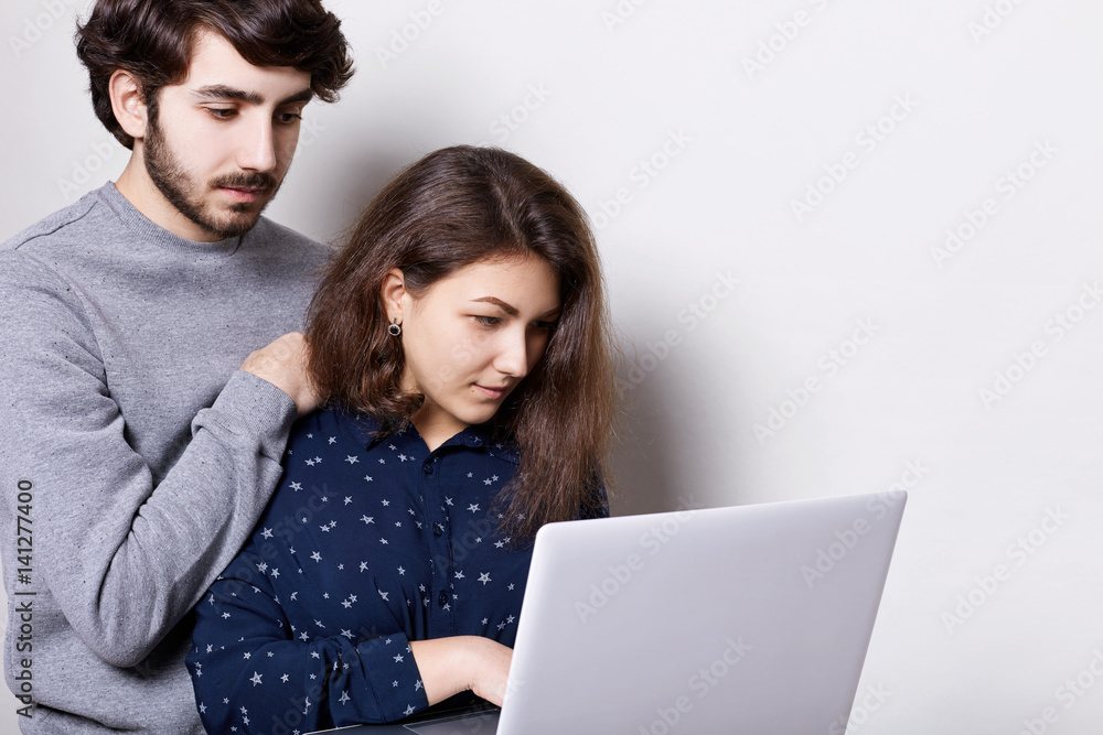 Young couple spending nice time together using high-speed internet connection on laptop. A man with trendy hairstyle and beard standing behind his girlfriend watching something on notebook Young couple spending nice time together using high-speed internet connection on laptop. A man with trendy hairstyle and beard standing behind his girlfriend watching something on notebook