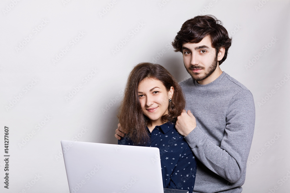 A happy man and woman standing sideways with laptop looking directly into camera isolated over white background. Stylish guy with beard standing behind his girlfriend enjoing modern technologies. A happy man and woman standing sideways with laptop looking directly into camera isolated over white background. Stylish guy with beard standing behind his girlfriend enjoing modern technologies.