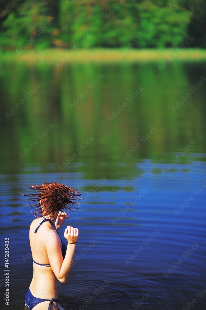 Fototapeta premium Young girl in bikini, standing in the river and shakes his head, flicking water out of your hair. Water drops fly in air, creating a beautiful pattern. Scenic summer background.