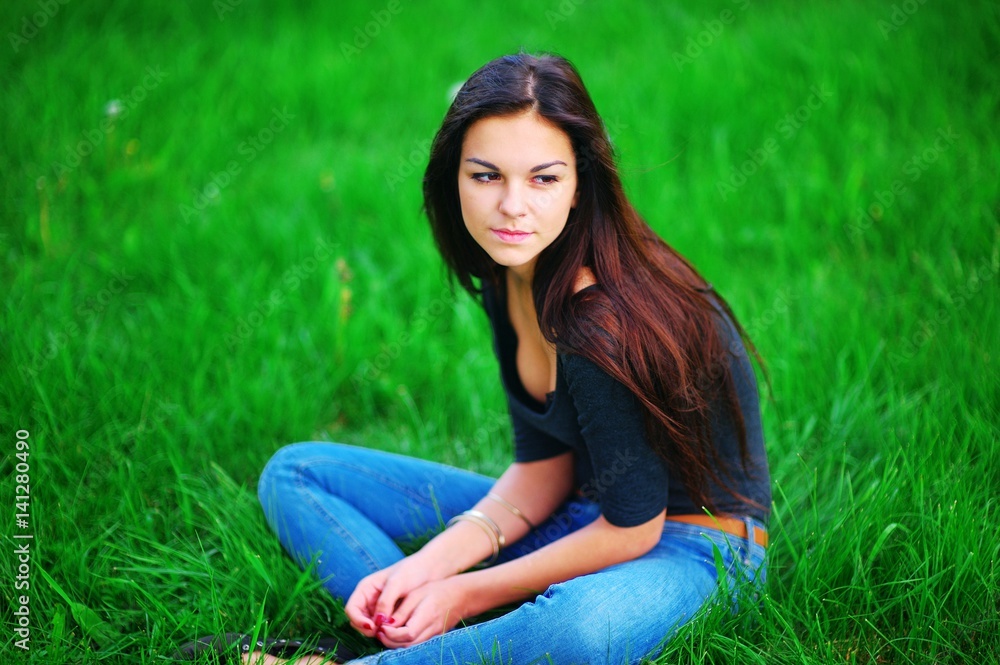 Portrait of young beautiful long-haired brooding girl in black shirt and blue jeans, sitting on the green lawn. Closeup. Portrait of young beautiful long-haired brooding girl in black shirt and blue jeans, sitting on the green lawn. Closeup.