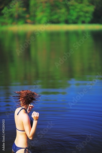 Fototapeta Naklejka Na Ścianę i Meble -  Young girl in bikini, standing in the river and shakes his head, flicking water out of your hair. Water drops fly in air, creating a beautiful pattern. Scenic summer background.