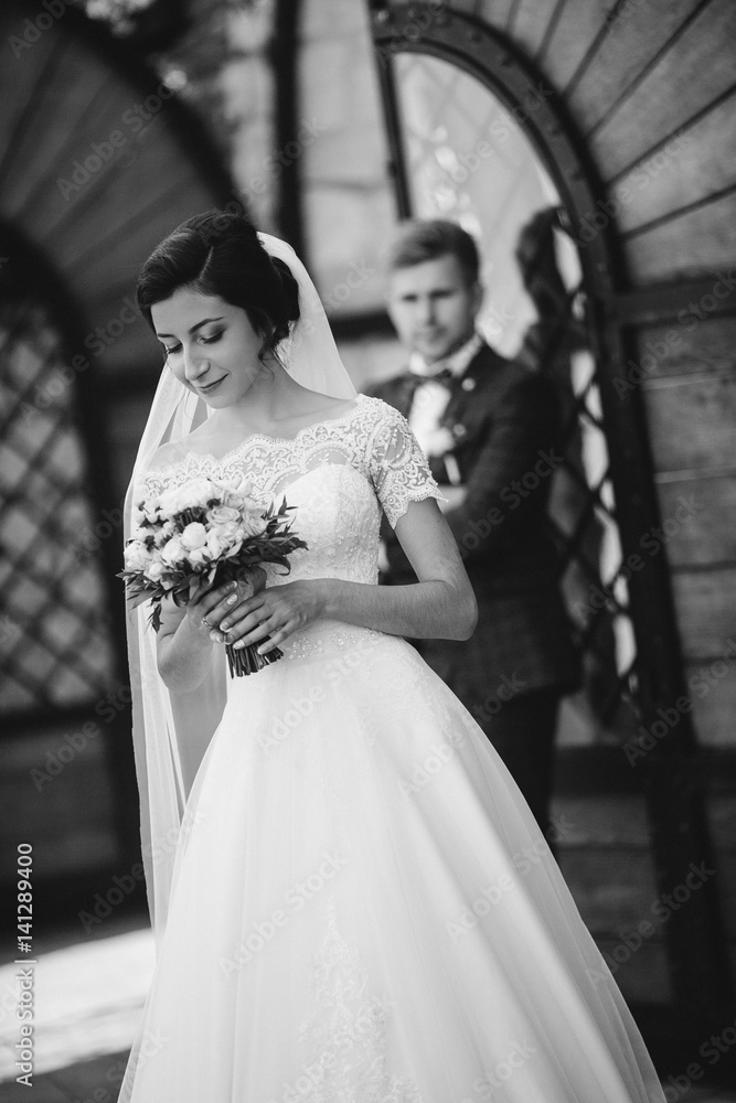 Handsome man groom hugging pretty girl or cute bride with beautiful brunette hair in white wedding dress outdoors at ancient building door on grey background