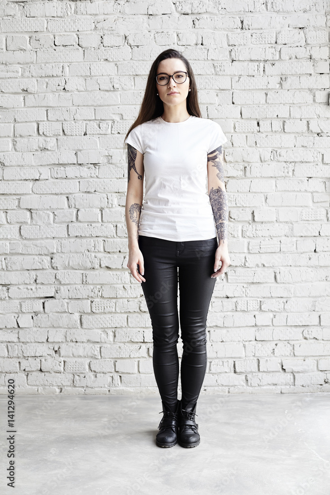 Young tattooed woman wearing blank t-shirt, standing in front of brick wall in loft Young tattooed woman wearing blank t-shirt, standing in front of brick wall in loft