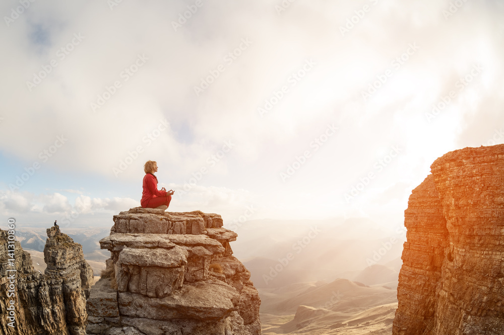 A beautiful girl meditates in a lotus pose sitting on a rock above the clouds against the backdrop of the sunset