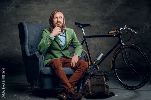 A man dressed in a green jacket sits on a chair with single speed bicycle on background.