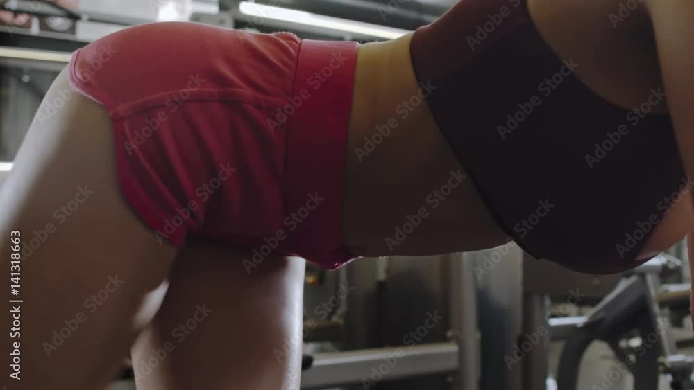 PAN of close up of seductive fit sportswoman in top and shorts standing in crawl position on flat bench in gym and doing leg lifts exercise 