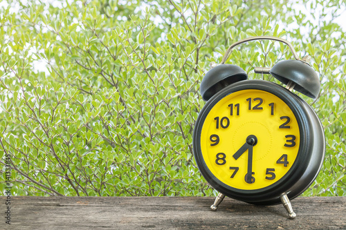 Closeup black and yellow alarm clock for decorate show half past seven or 7:30 a.m. on old brown wood desk on green leaves in the park textured background